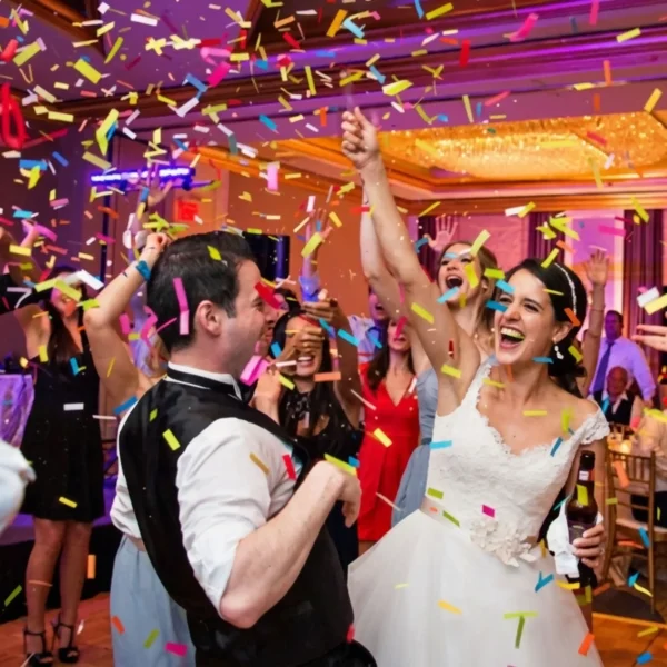 A joyful bride and groom dancing on a packed wedding reception dance floor surrounded by falling multi-coloured confetti and cheered on by guests.